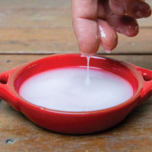Load image into Gallery viewer, A close-up of a hand squeezing our white coloured pure emu oil showing it's consistency, with the liquid dripping into a small red ceramic bowl on a wooden surface.