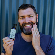 Load image into Gallery viewer, Middle aged man holding our 100ml pure emu oil in one hand and applying it to his beard in the other hand with blue corrugated iron background
